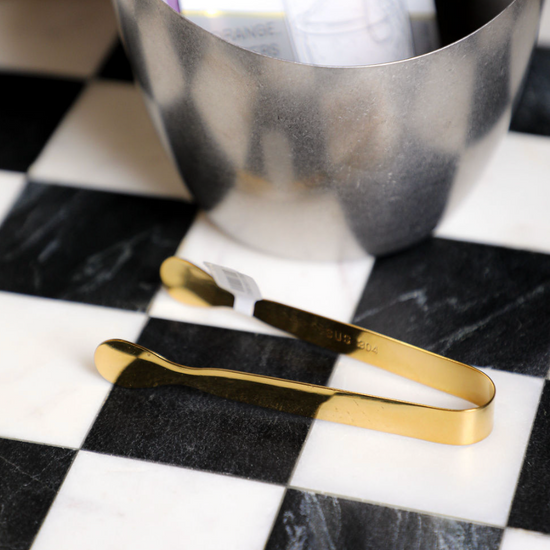 Gold-colored stainless steel ice tongs by unknown vendor, on a black and white checkered surface, next to a metal container.