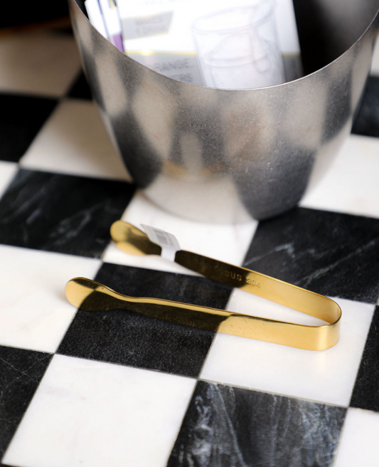 Gold-colored stainless steel ice tongs by unknown vendor, on a black and white checkered surface, next to a metal container.