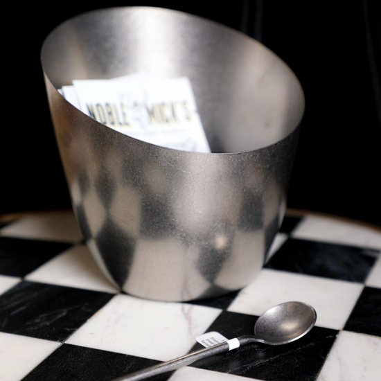 Stainless steel slanted container by Noble Mick's, holding white papers, sitting on a black and white checkered surface with a long spoon.