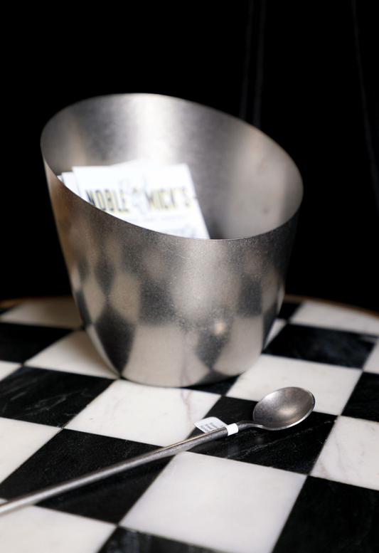 Stainless steel slanted container by Noble Mick's, holding white papers, sitting on a black and white checkered surface with a long spoon.
