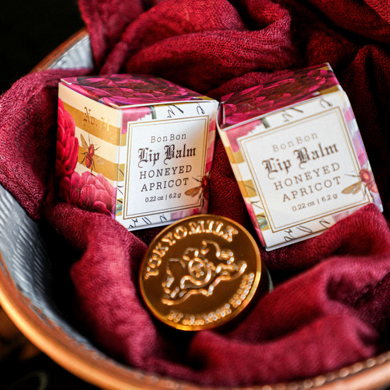 Honeyed apricot lip balm by TokyoMilk, styled with a golden token and a red gauze-like fabric in a speckled metal bowl.
