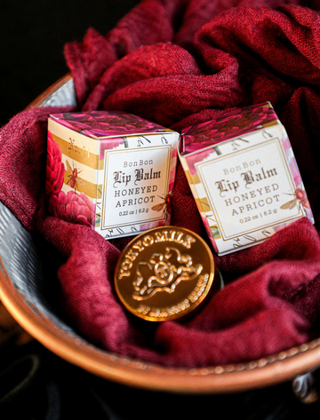 Honeyed apricot lip balm by TokyoMilk, styled with a golden token and a red gauze-like fabric in a speckled metal bowl.

