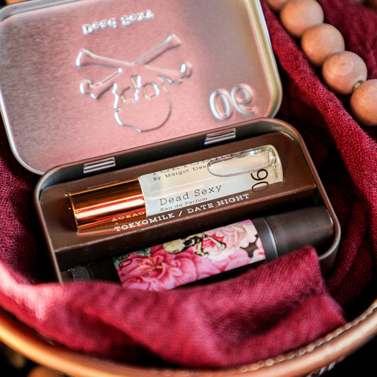 Bath and body kit by Tokyo Milk, including perfume and balm, contained within a decorative metal tin, nestled among ruddy fabric and wooden beads.
