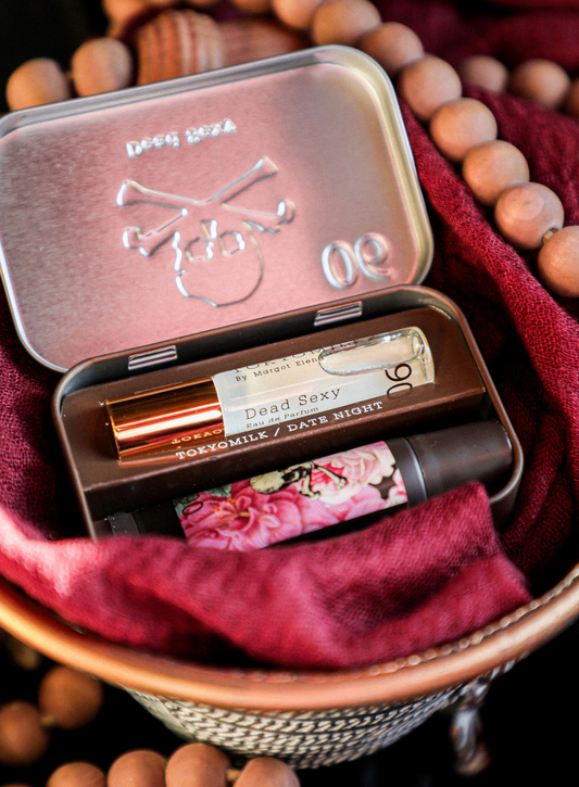Bath and body kit by Tokyo Milk, including perfume and balm, contained within a decorative metal tin, nestled among ruddy fabric and wooden beads.
