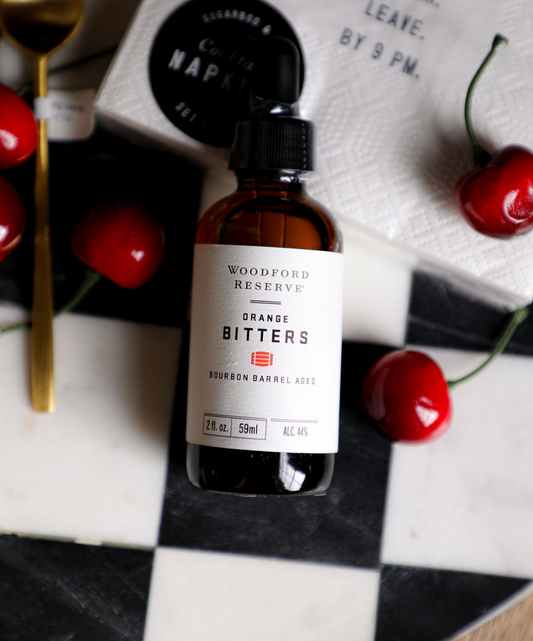 Bottle of orange bitters by Woodford Reserve, displayed with cocktail napkins and cherries on a checkered surface.