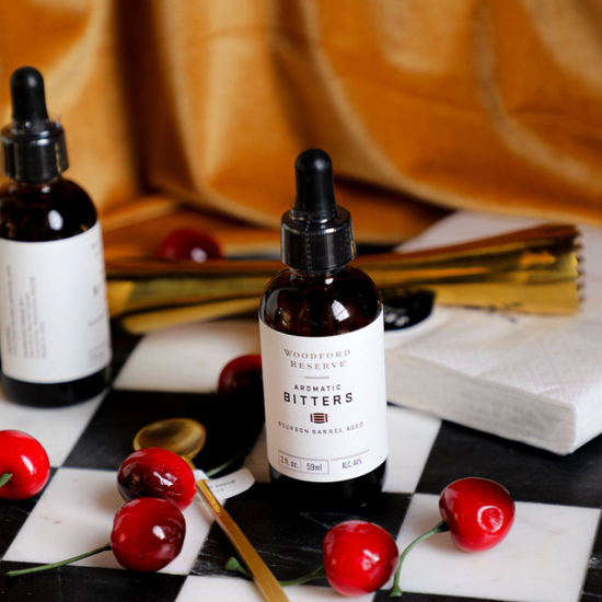 Aromatic bitters by Woodford Reserve, alongside decorative cherries, on a black and white checkerboard surface.