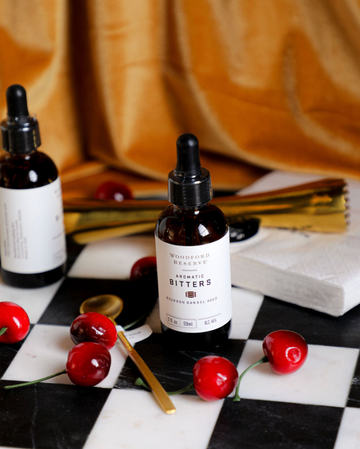 Aromatic bitters by Woodford Reserve, alongside decorative cherries, on a black and white checkerboard surface.