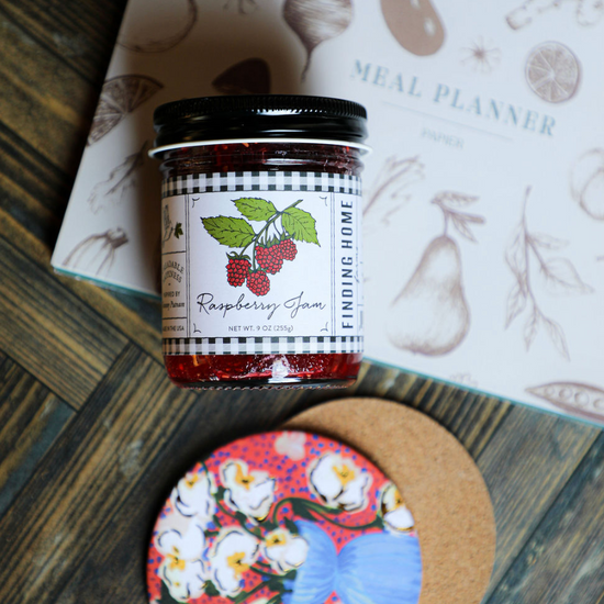 Jar of raspberry jam by Finding Home, with a black and white checkered label, on a colorful floral coaster.
