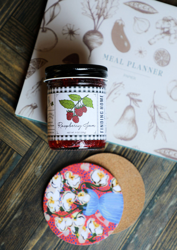 Jar of raspberry jam by Finding Home, with a black and white checkered label, on a colorful floral coaster.
