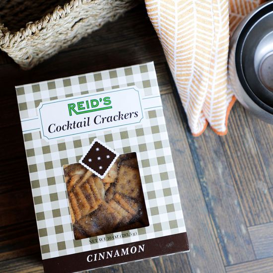 Box of cinnamon cocktail crackers by Reid's, displayed on a wood surface near a woven basket and patterned orange towel.