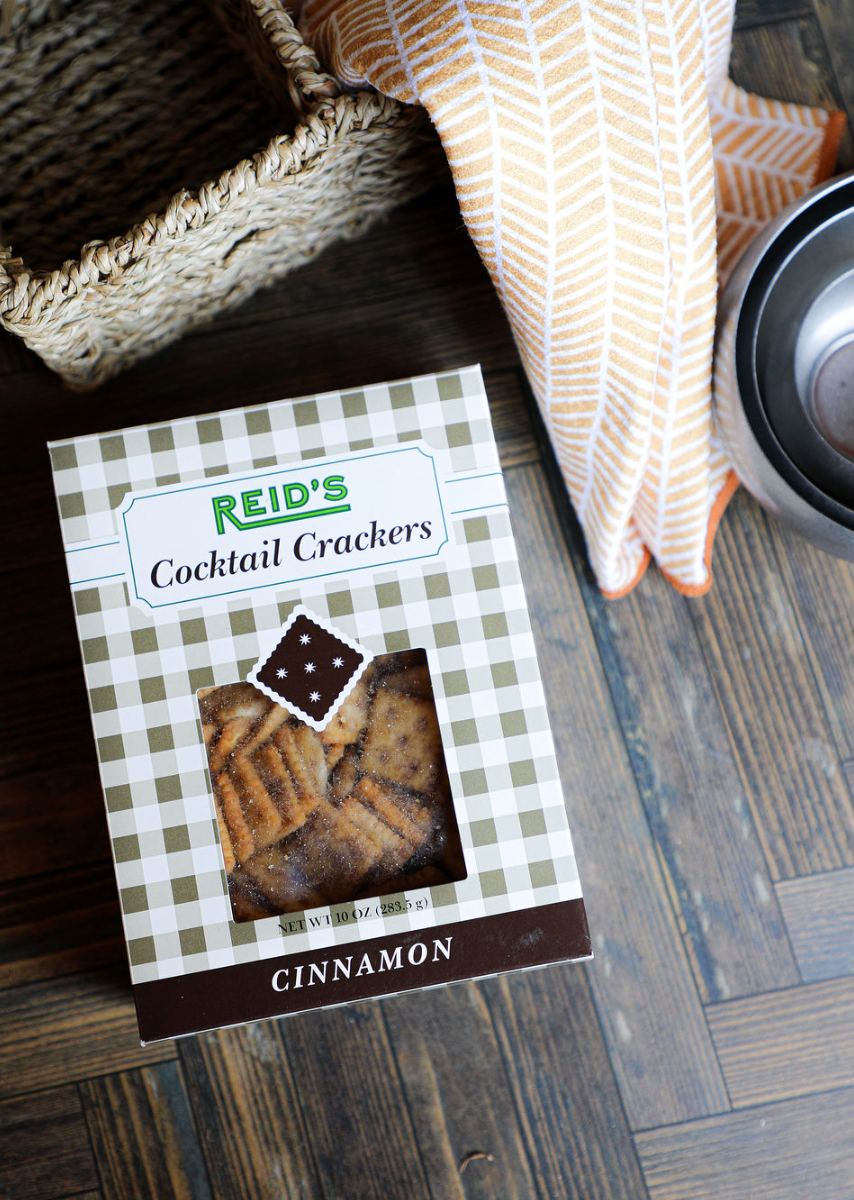 Box of cinnamon cocktail crackers by Reid's, displayed on a wood surface near a woven basket and patterned orange towel.