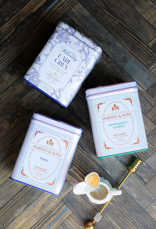 Three tea tins by Harney & Sons, with Earl Grey, Paris, and Peppermint Herbal varieties, sitting on a wooden surface.