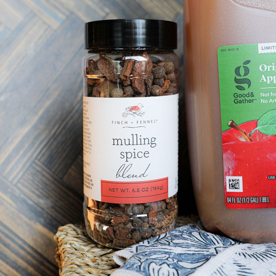 Mulling spice blend by Finch & Fennel, in a clear jar, next to apple cider.
