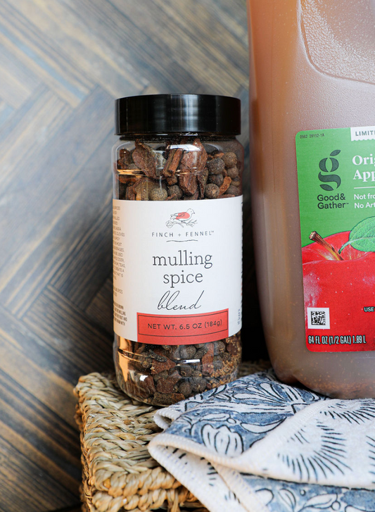 Mulling spice blend by Finch & Fennel, in a clear jar, next to apple cider.
