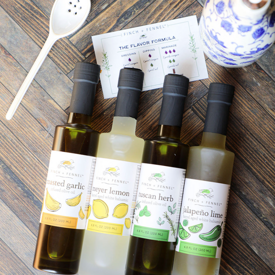 Gourmet barrel aged balsamics and infused olive oils from Finch + Fennel, arranged on a wooden surface with a white spoon and recipe card.
