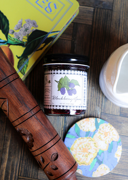 Jar of Blackberry Jam by Finding Home, on a wooden table with a rolling pin and a yellow box.
