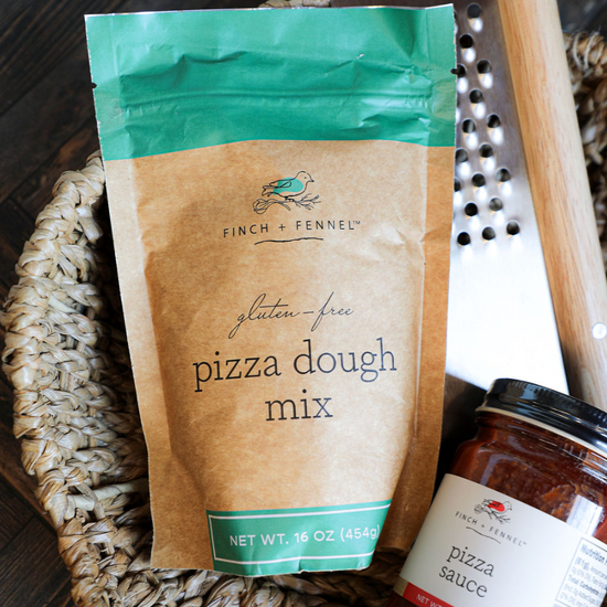 Gluten-free pizza dough mix from Finch + Fennel, next to pizza sauce and a grater in a woven basket.
