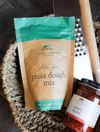 Gluten-free pizza dough mix from Finch + Fennel, next to pizza sauce and a grater in a woven basket.
