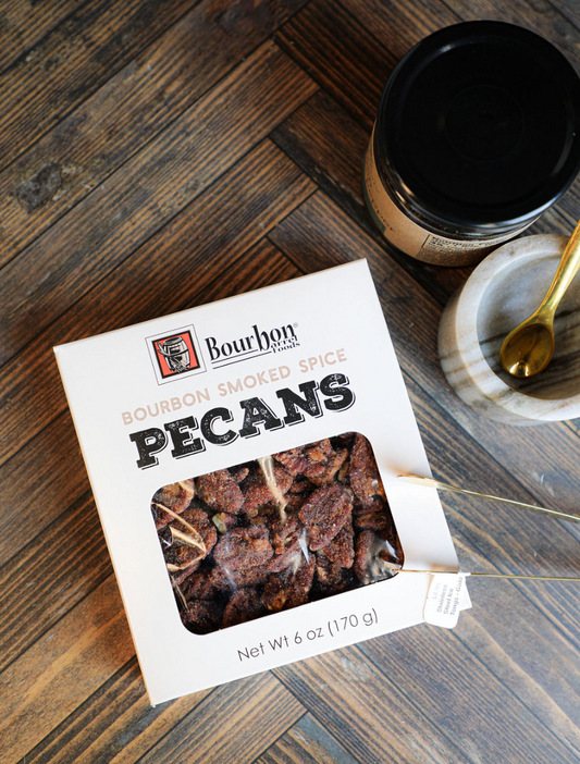 Bourbon smoked spiced pecans by Bourbon Barrel Foods with stainless steel ice tongs on wooden herringbone background.
