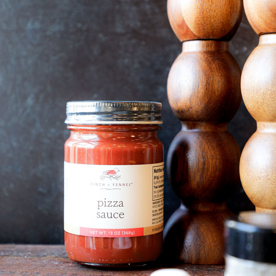 Jar of pizza sauce by Finch + Fennel, sitting on a wooden surface next to wooden spice mills.