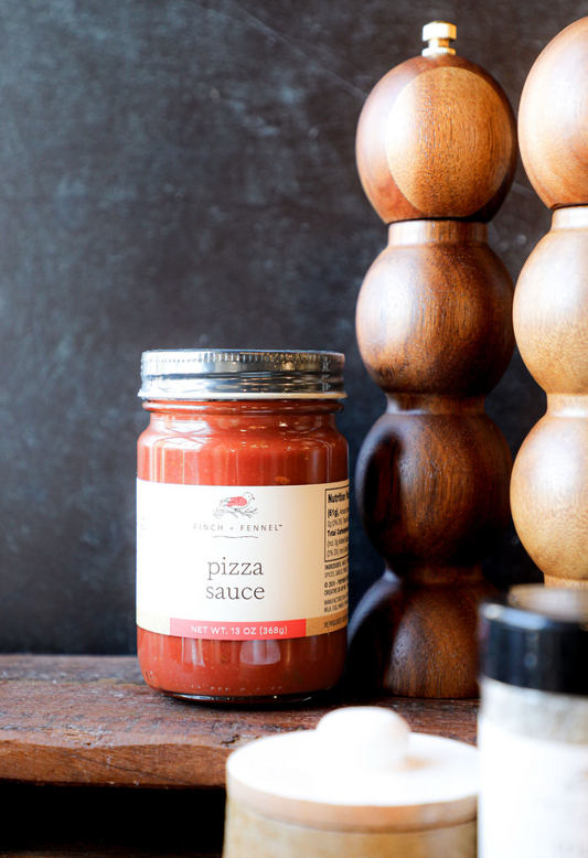 Jar of pizza sauce by Finch + Fennel, sitting on a wooden surface next to wooden spice mills.