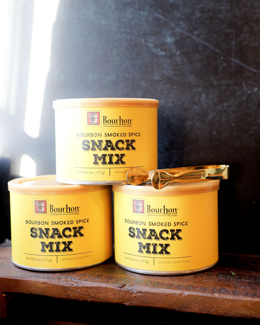 Yellow snack mix tins by Bourbon Barrel Foods, stacked with golden tongs resting on top, on a wooden surface.