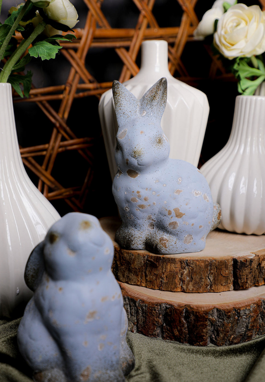 Two blue ceramic rabbit figurines on a wooden surface with white vases and flowers in the background.