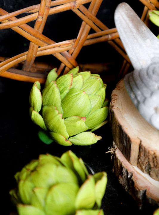 Four inch green faux artichoke, surrounded by wicker and a wood plank.