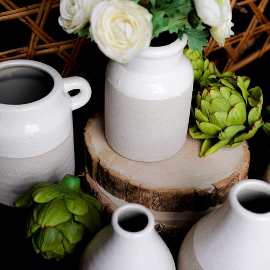 Ceramic two-tone glazed pots with a cream and beige color scheme, surrounded by faux artichokes and set on a wood slice.