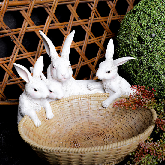 Three white rabbit figurines in a woven basket with a wicker wall and greenery in the background.
