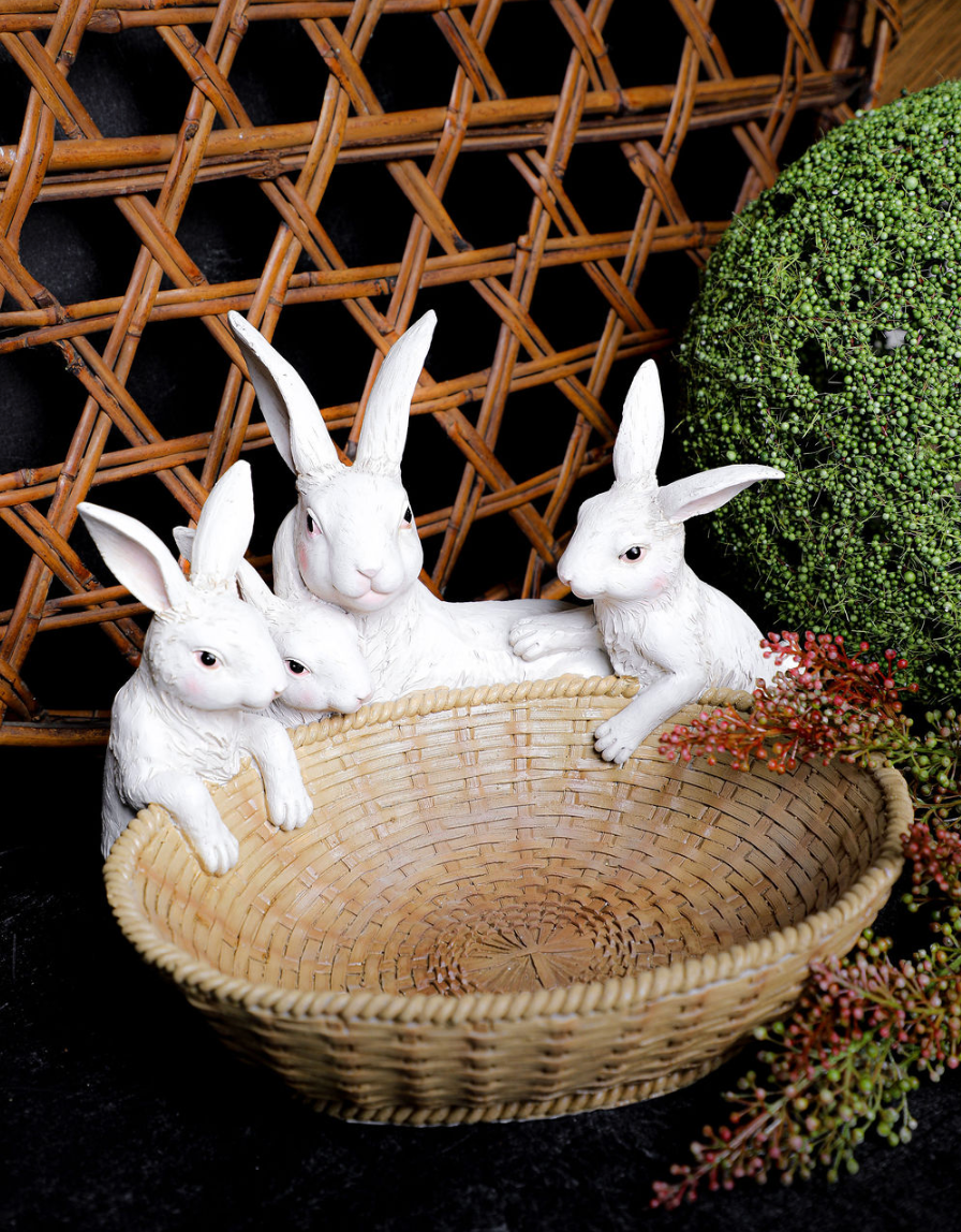 Three white rabbit figurines in a woven basket with a wicker wall and greenery in the background.
