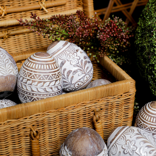 Whitewashed floral resin Easter egg with carved designs, in a woven basket with decorative foliage.
