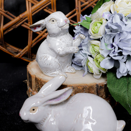 Two white ceramic rabbit figurines on a wooden block with flowers in the background.