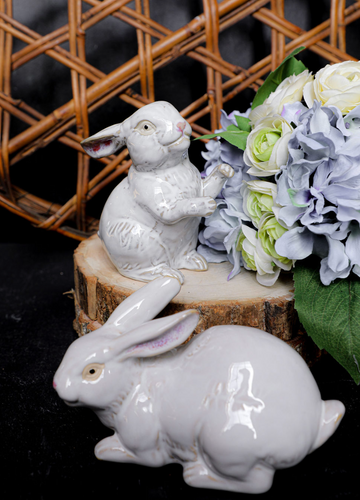 Two white ceramic rabbit figurines on a wooden block with flowers in the background.