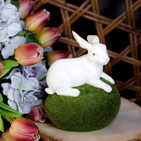 Decorative white rabbit figurine on a moss egg atop a wooden slice, surrounded by faux flowers.