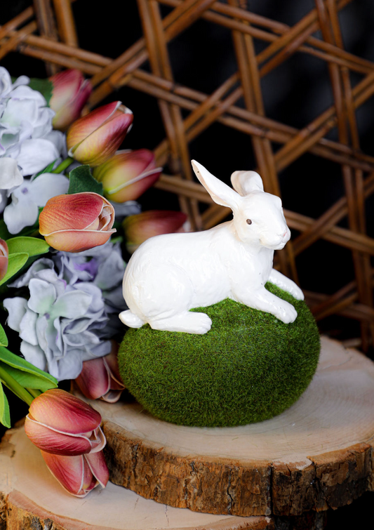 Decorative white rabbit figurine on a moss egg atop a wooden slice, surrounded by faux flowers.
