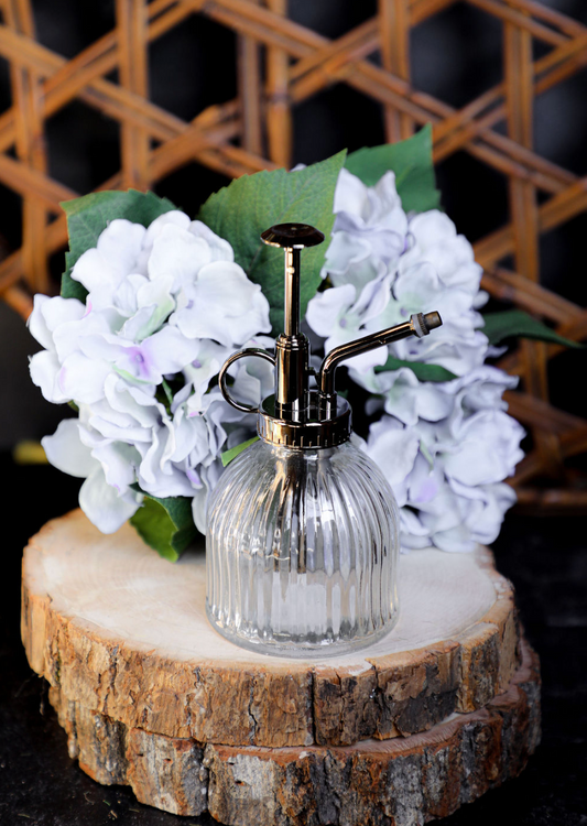 Clear glass mister with metal pump, surrounded by flowers, on a wood slice.