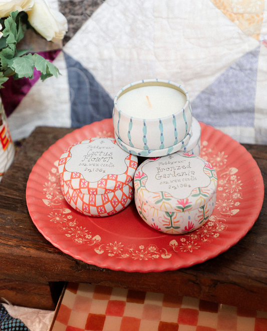 Three small candles with decorative lids on a pink plate with floral patterns, placed on a wooden surface.
