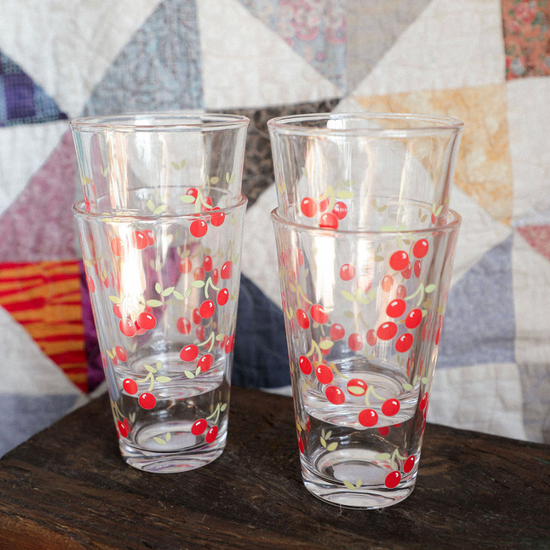 Two glass tumblers with cherry design on a wooden surface in front of a quilted fabric background.