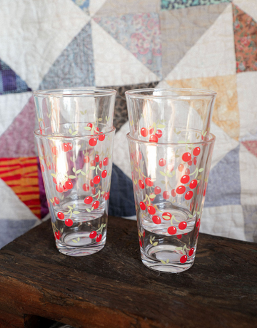 Two glass tumblers with cherry design on a wooden surface in front of a quilted fabric background.
