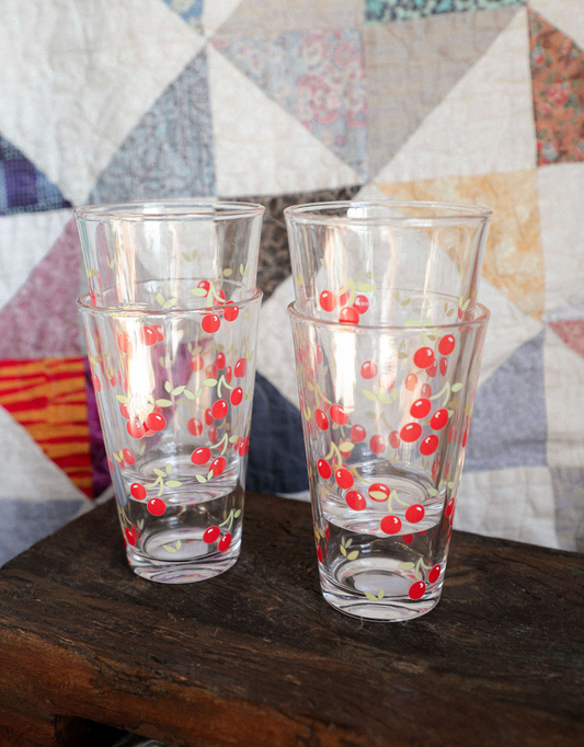 Two glass tumblers with cherry design on a wooden surface in front of a quilted fabric background.