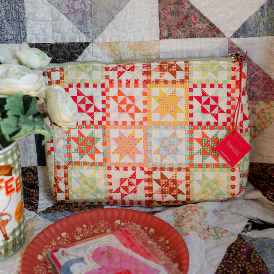 Colorful patchwork pouch on a quilt with a red plate and floral arrangement.