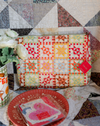 Colorful patchwork pouch on a quilt with a red plate and floral arrangement.
