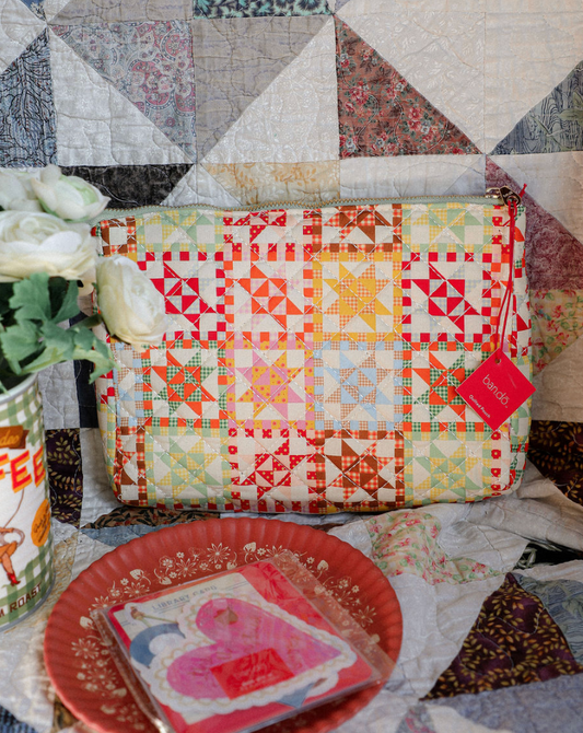 Colorful patchwork pouch on a quilt with a red plate and floral arrangement.