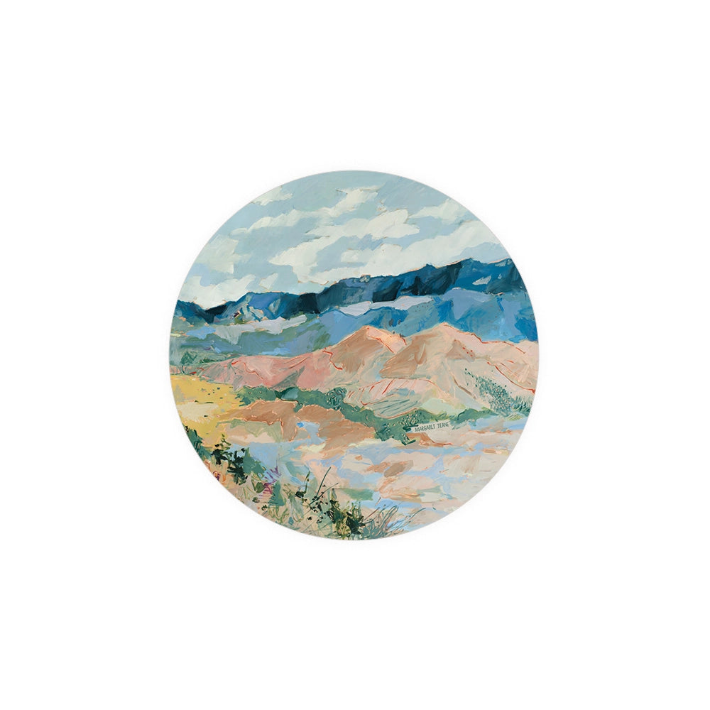 Round landscape art coaster by Margaret Teane, featuring abstract mountains, desert landscape, and sky with clouds.
